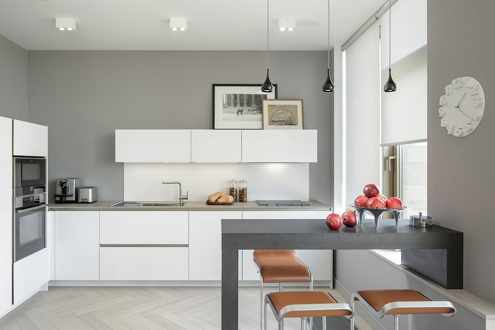 Modern inredning av ett kök, med en undermonterad diskho, vita skåp, laminatbänkskiva, vitt stänkskydd, svarta vitvaror, beiget golv, släta luckor, ljust trägolv och en halv köksö