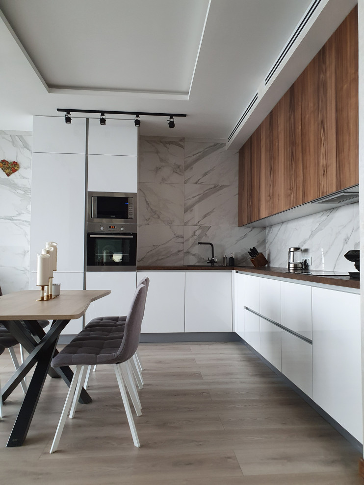 Photo of a medium sized contemporary l-shaped kitchen/diner in Other with a built-in sink, flat-panel cabinets, white cabinets, wood worktops, grey splashback, porcelain splashback, stainless steel appliances, no island, brown floors and brown worktops.