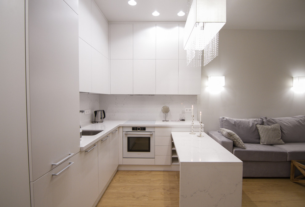 Foto på ett mellanstort funkis vit kök, med en enkel diskho, släta luckor, vita skåp, bänkskiva i kvarts, vitt stänkskydd, stänkskydd i sten, vita vitvaror, ljust trägolv, en halv köksö och brunt golv