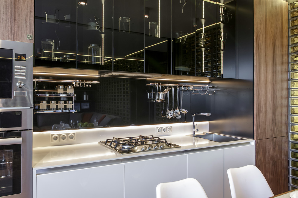 Стильный дизайн: прямая кухня-гостиная среднего размера в современном стиле с одинарной мойкой, плоскими фасадами, черным фартуком, фартуком из стекла, техникой из нержавеющей стали и белой столешницей - последний тренд