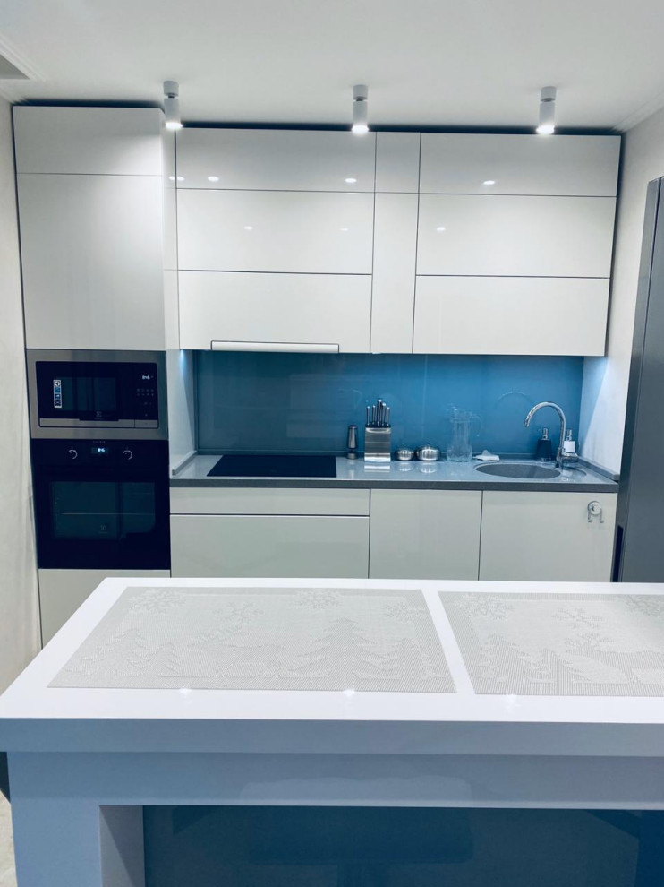 This is an example of a small contemporary single-wall kitchen/diner in Other with a submerged sink, flat-panel cabinets, white cabinets, composite countertops, blue splashback, glass sheet splashback, black appliances, porcelain flooring, an island, white floors and grey worktops.