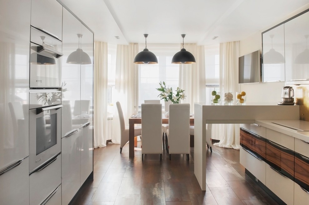 На фото: параллельная кухня в современном стиле с обеденным столом, плоскими фасадами, белой техникой, полуостровом, коричневым полом, белыми фасадами и фартуком цвета металлик с