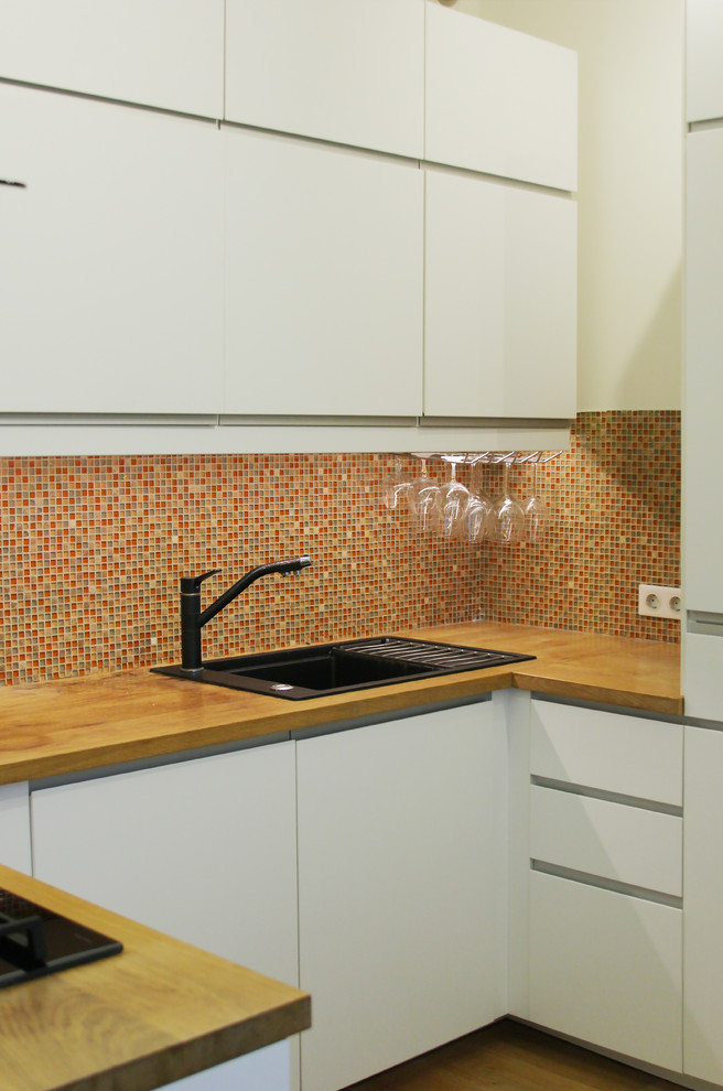 Inredning av ett minimalistiskt litet kök, med en nedsänkt diskho, släta luckor, vita skåp, träbänkskiva, orange stänkskydd, stänkskydd i mosaik, rostfria vitvaror och mellanmörkt trägolv