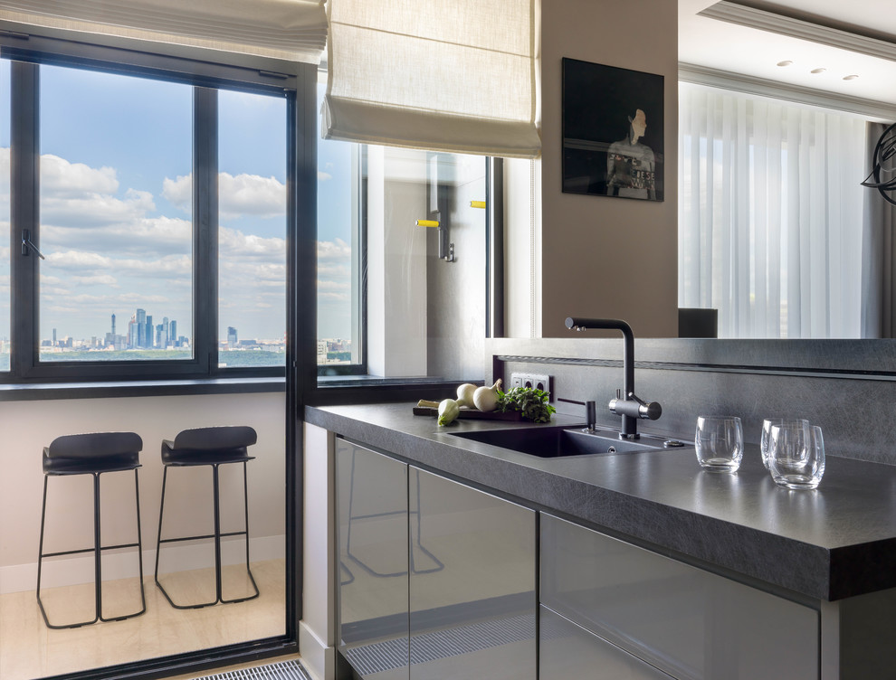 Medium sized contemporary single-wall open plan kitchen in Other with flat-panel cabinets, grey cabinets, grey splashback, porcelain splashback, marble flooring, a breakfast bar, beige floors, black worktops, a built-in sink and granite worktops.