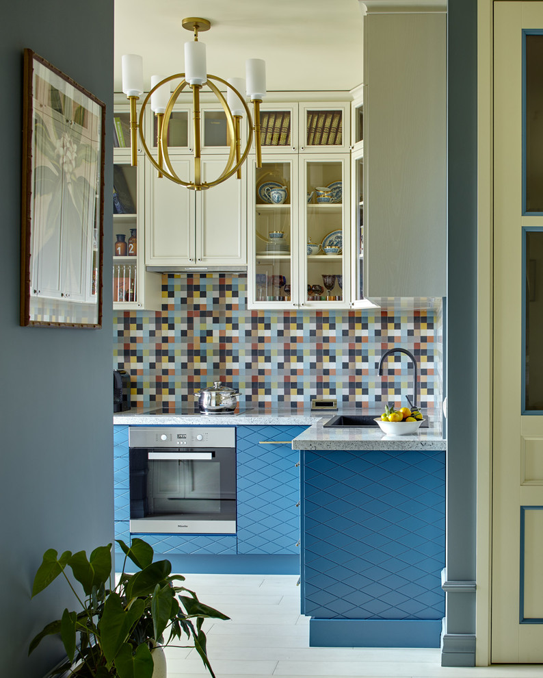 Идея дизайна: угловая кухня-гостиная в стиле неоклассика (современная классика) с накладной мойкой, плоскими фасадами, синими фасадами, разноцветным фартуком, техникой из нержавеющей стали и белым полом без острова