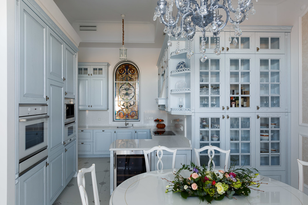 На фото: п-образная кухня в классическом стиле с обеденным столом, врезной мойкой, фасадами с выступающей филенкой, синими фасадами, белым фартуком, белой техникой и полуостровом с