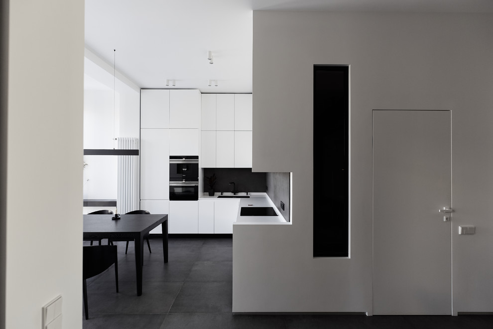 Пример оригинального дизайна: угловая кухня в современном стиле с обеденным столом, врезной мойкой, плоскими фасадами, черным фартуком, черной техникой, черным полом, белой столешницей и черно-белыми фасадами