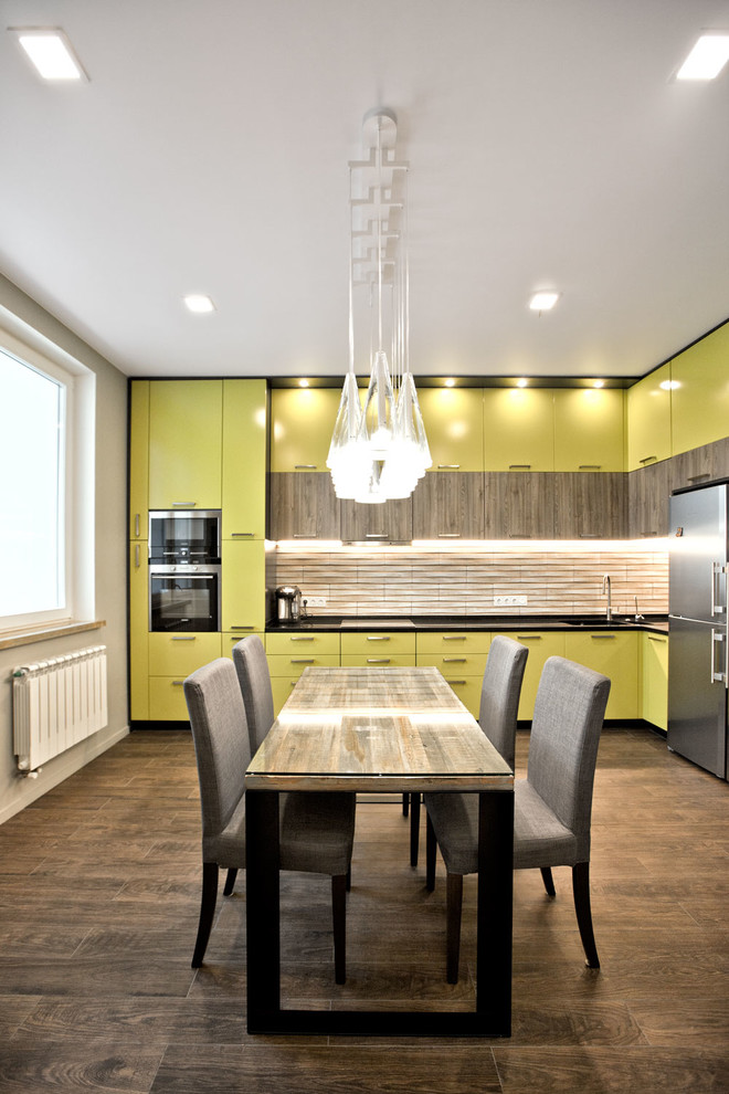 Свежая идея для дизайна: угловая кухня в современном стиле с обеденным столом, плоскими фасадами, желтыми фасадами, бежевым фартуком, техникой из нержавеющей стали и коричневым полом - отличное фото интерьера
