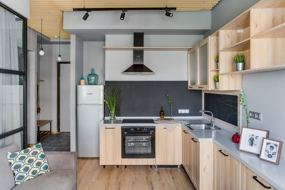 Идея дизайна: угловая, светлая кухня-гостиная в скандинавском стиле с накладной мойкой, светлыми деревянными фасадами, черным фартуком, черной техникой, светлым паркетным полом, серой столешницей и бежевым полом без острова