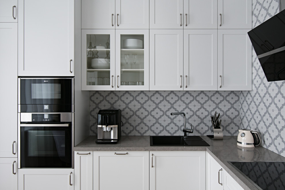 Foto på ett funkis l-kök, med luckor med infälld panel, vita skåp, grått stänkskydd och svarta vitvaror
