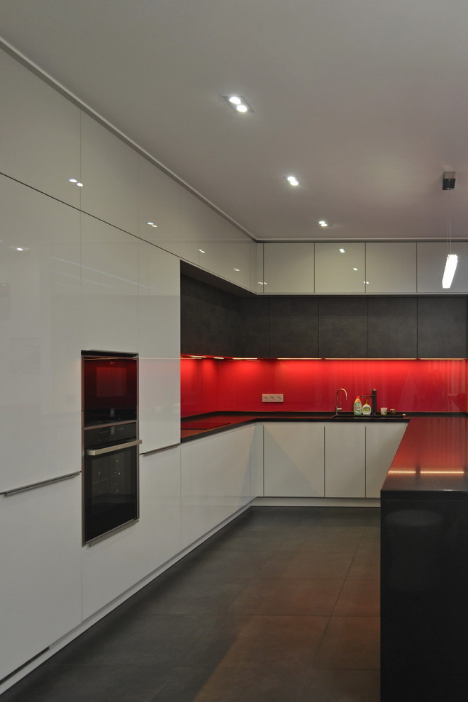 Modern inredning av ett stort svart svart kök, med en undermonterad diskho, släta luckor, vita skåp, bänkskiva i kvarts, rött stänkskydd, stänkskydd i glaskakel, svarta vitvaror, klinkergolv i porslin, en halv köksö och svart golv
