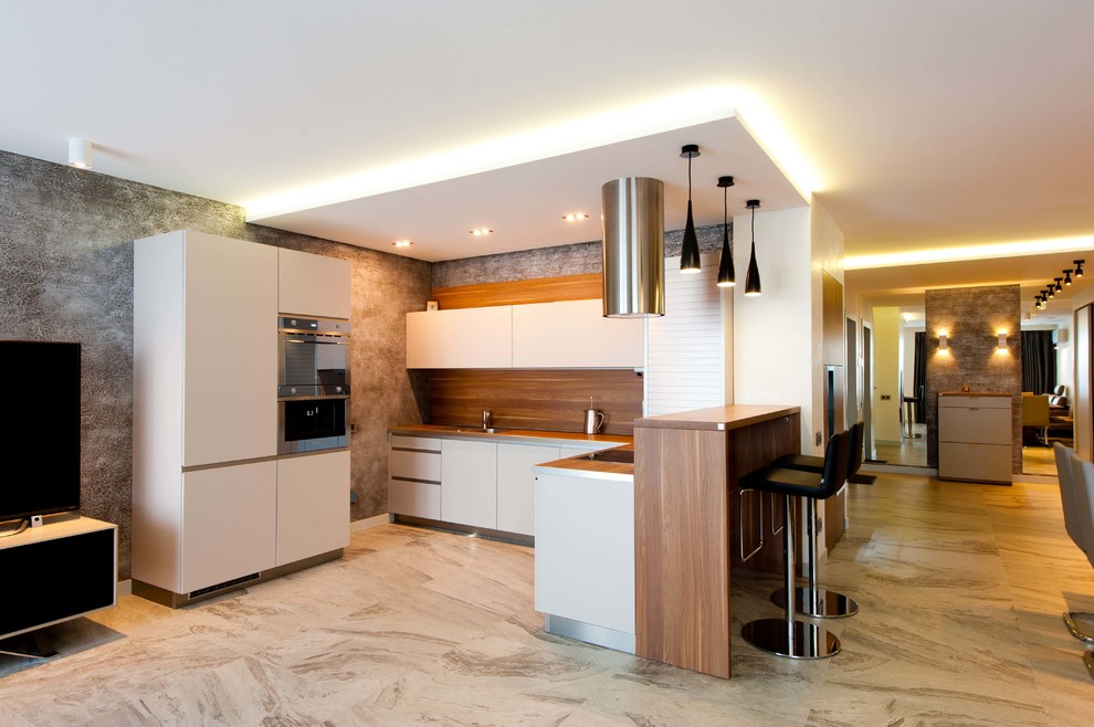 Modern inredning av ett kök, med släta luckor, vita skåp, brunt stänkskydd, rostfria vitvaror och en halv köksö