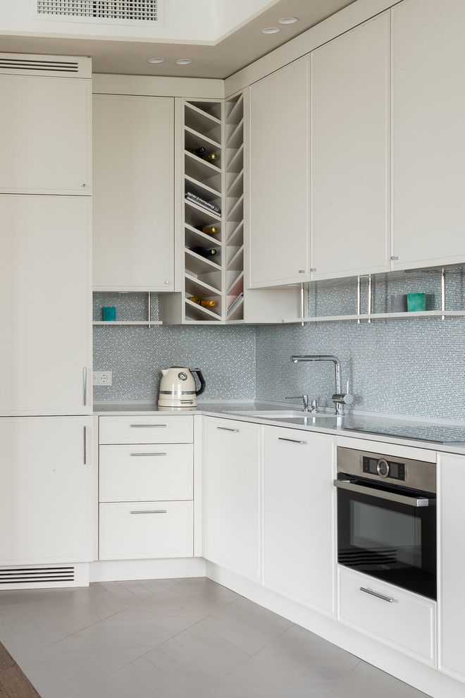 Bild på ett funkis vit vitt l-kök, med en undermonterad diskho, luckor med profilerade fronter, vita skåp, grått stänkskydd, rostfria vitvaror och grått golv