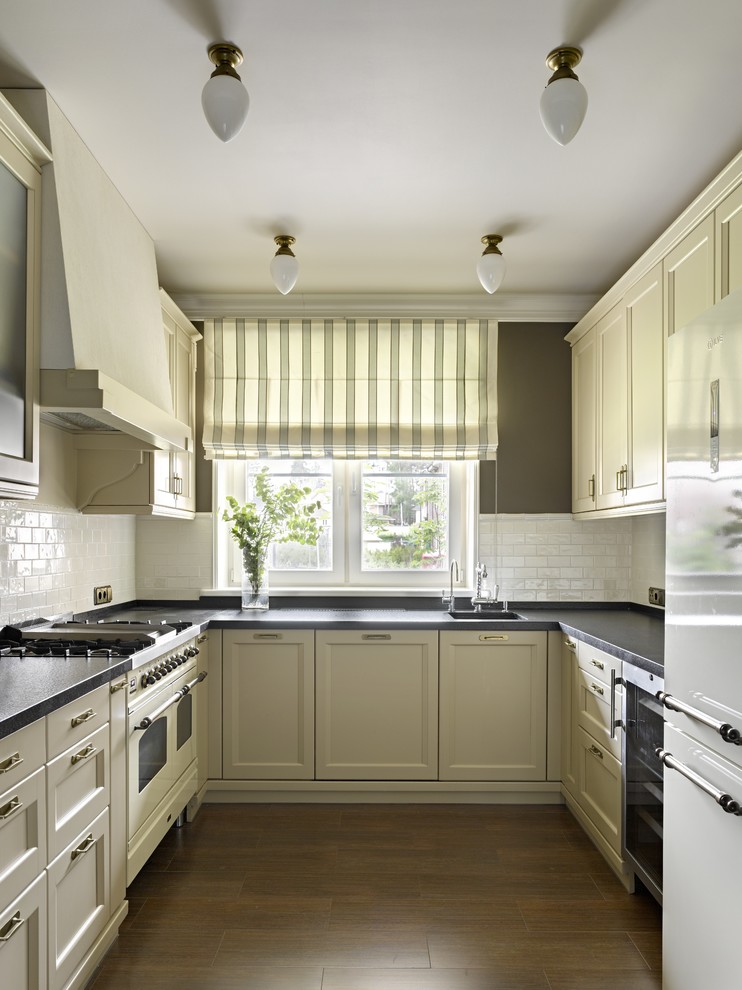 Exempel på ett avskilt, stort modernt u-kök, med en enkel diskho, luckor med infälld panel, vita vitvaror, mellanmörkt trägolv, fönster som stänkskydd, beige skåp och vitt stänkskydd