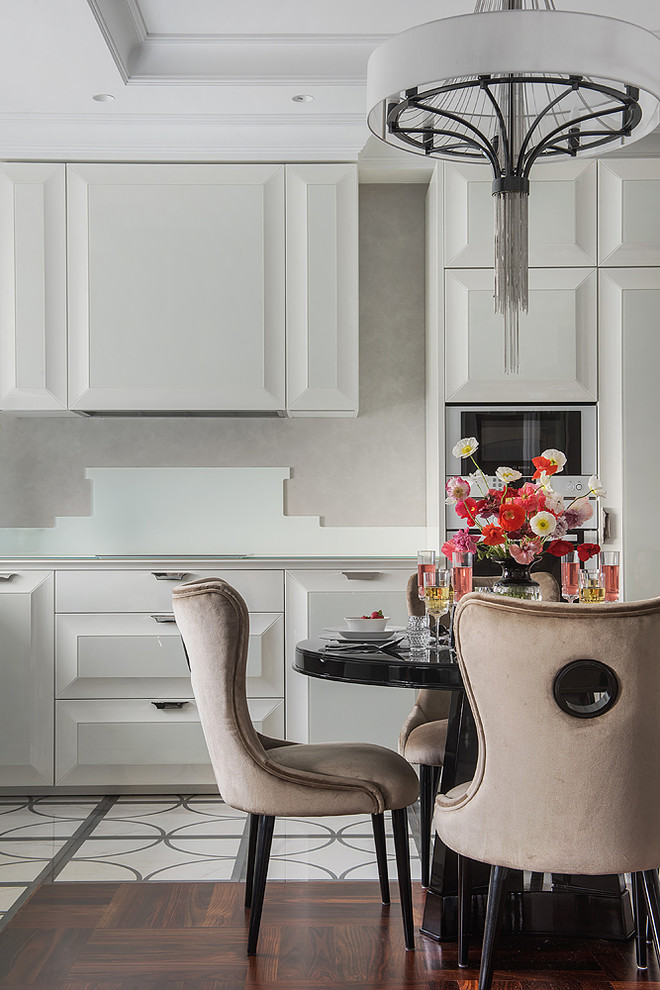 На фото: угловая кухня в стиле неоклассика (современная классика) с обеденным столом, фасадами с выступающей филенкой, серым фартуком, белыми фасадами и белой техникой