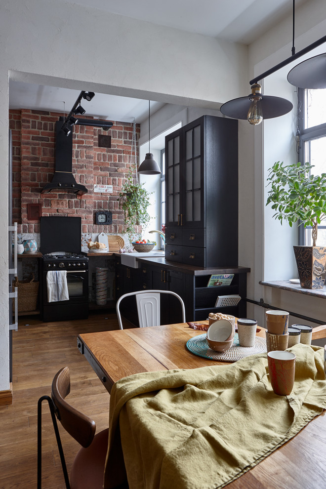 Example of an urban l-shaped medium tone wood floor and brown floor eat-in kitchen design in Moscow with a farmhouse sink, shaker cabinets, black cabinets, wood countertops, black appliances, no island and brown countertops