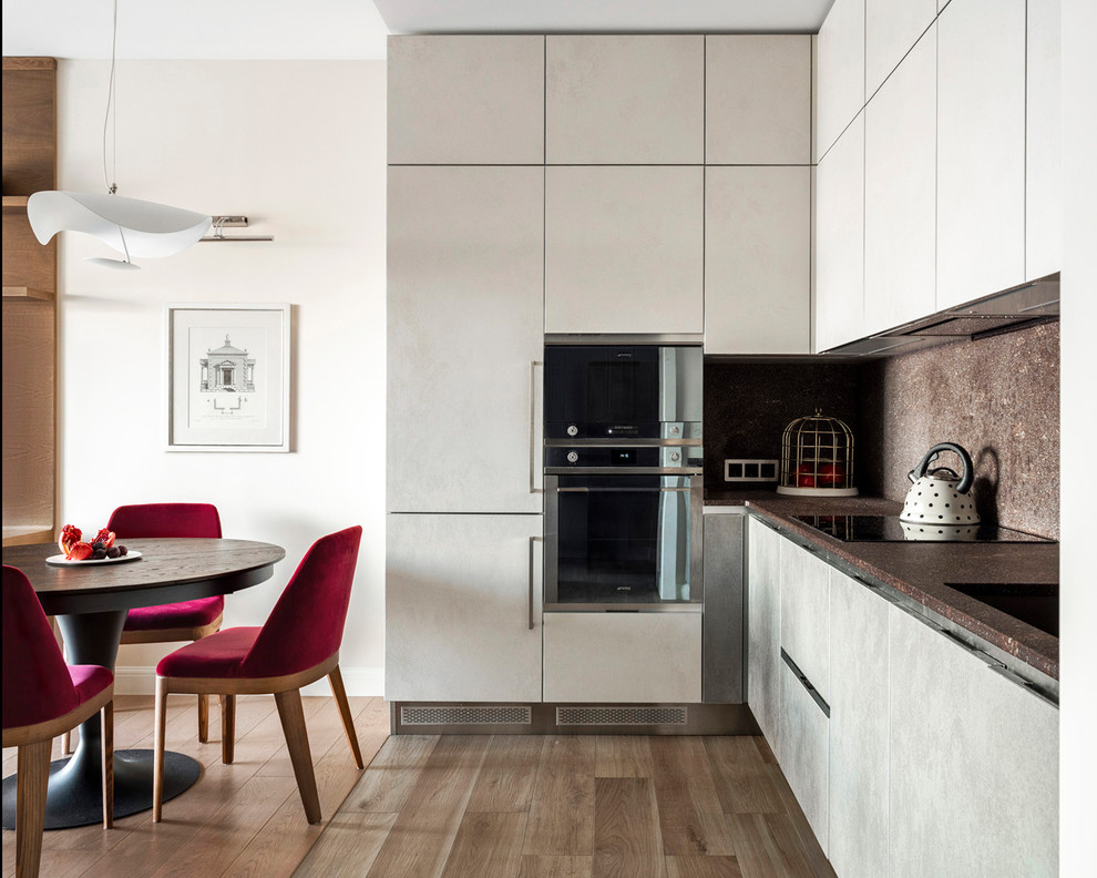 Стильный дизайн: угловая кухня в современном стиле с обеденным столом, плоскими фасадами, серыми фасадами, черной техникой, паркетным полом среднего тона и коричневым полом - последний тренд