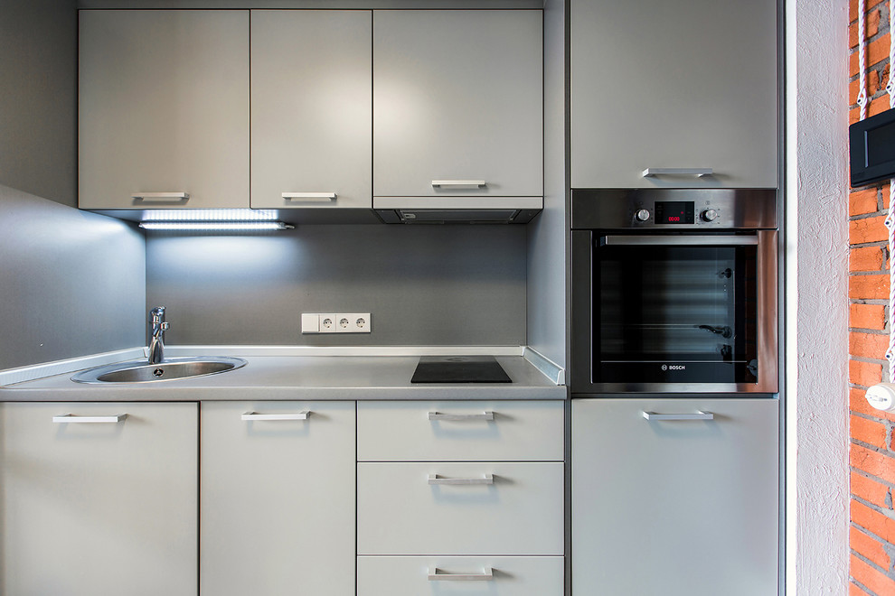 На фото: прямая кухня-гостиная в современном стиле с накладной мойкой, плоскими фасадами, серыми фасадами, серым фартуком и техникой из нержавеющей стали без острова
