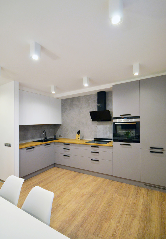 На фото: угловая кухня в современном стиле с накладной мойкой, плоскими фасадами, серыми фасадами, деревянной столешницей, серым фартуком, светлым паркетным полом, коричневым полом и коричневой столешницей