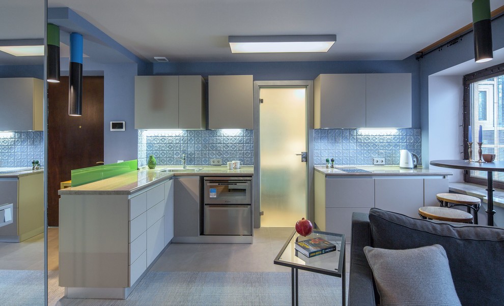 Стильный дизайн: угловая кухня-гостиная в современном стиле с одинарной мойкой, плоскими фасадами, белыми фасадами, синим фартуком, техникой из нержавеющей стали и серым полом без острова - последний тренд
