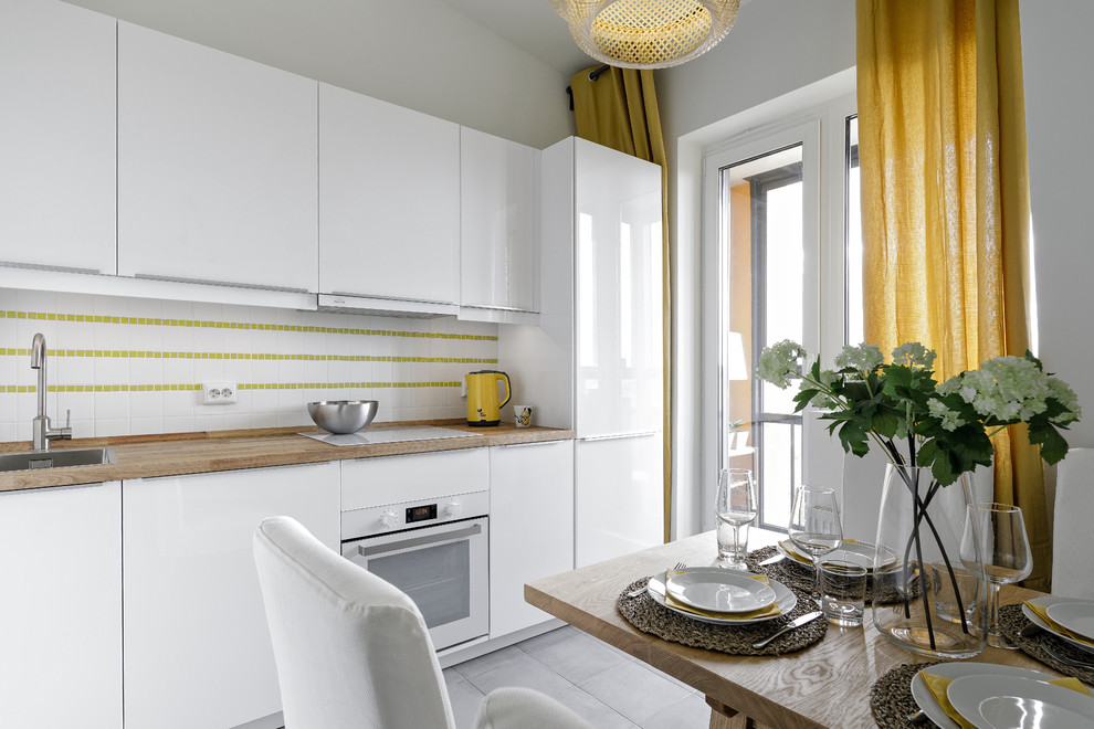 Example of a mid-sized danish single-wall ceramic tile and gray floor eat-in kitchen design in Saint Petersburg with a drop-in sink, flat-panel cabinets, white cabinets, wood countertops, ceramic backsplash, white appliances and no island
