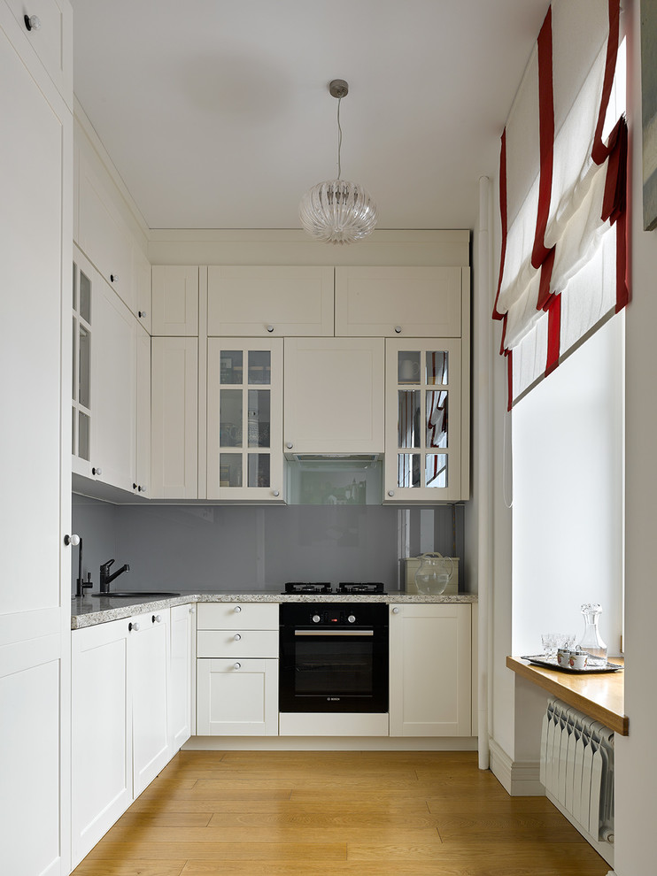 Bild på ett avskilt vintage grå grått l-kök, med en nedsänkt diskho, vita skåp, bänkskiva i kvartsit, glaspanel som stänkskydd, svarta vitvaror, grått stänkskydd, mellanmörkt trägolv, brunt golv och skåp i shakerstil