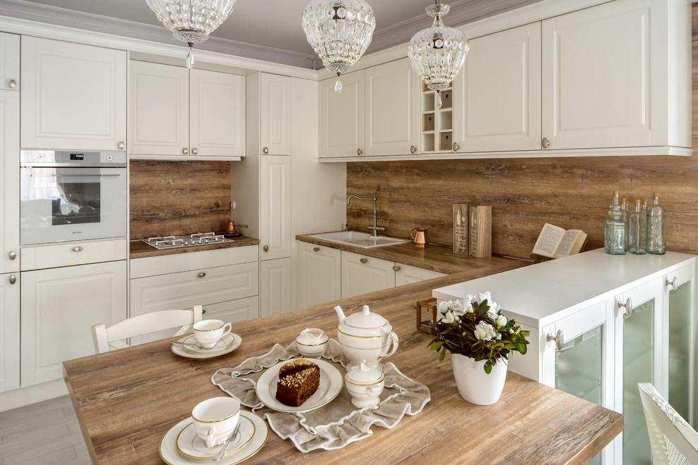 Inspiration for a timeless u-shaped eat-in kitchen remodel in Other with a drop-in sink, raised-panel cabinets, wood countertops, white appliances, a peninsula, white cabinets and brown backsplash