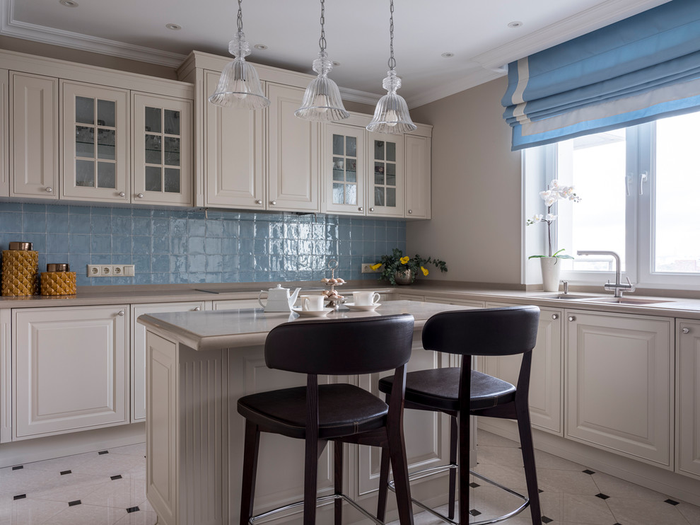 На фото: угловая кухня в стиле неоклассика (современная классика) с фасадами с выступающей филенкой, белыми фасадами, синим фартуком, островом, белым полом и бежевой столешницей с