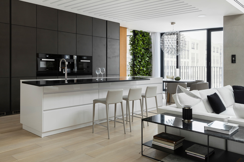 Foto de cocina contemporánea abierta con fregadero integrado, electrodomésticos negros, una isla, armarios con paneles lisos, suelo de madera clara, encimeras negras y con blanco y negro