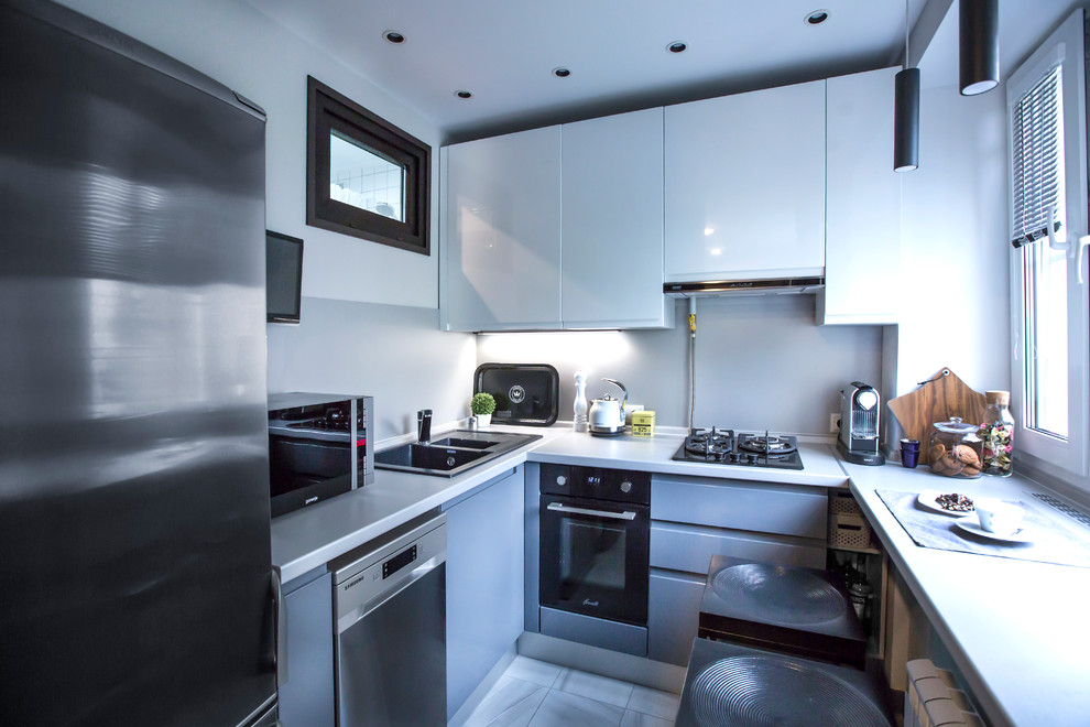 Идея дизайна: угловая кухня-гостиная в современном стиле с накладной мойкой, плоскими фасадами, белыми фасадами, белым фартуком, черной техникой и серым полом без острова
