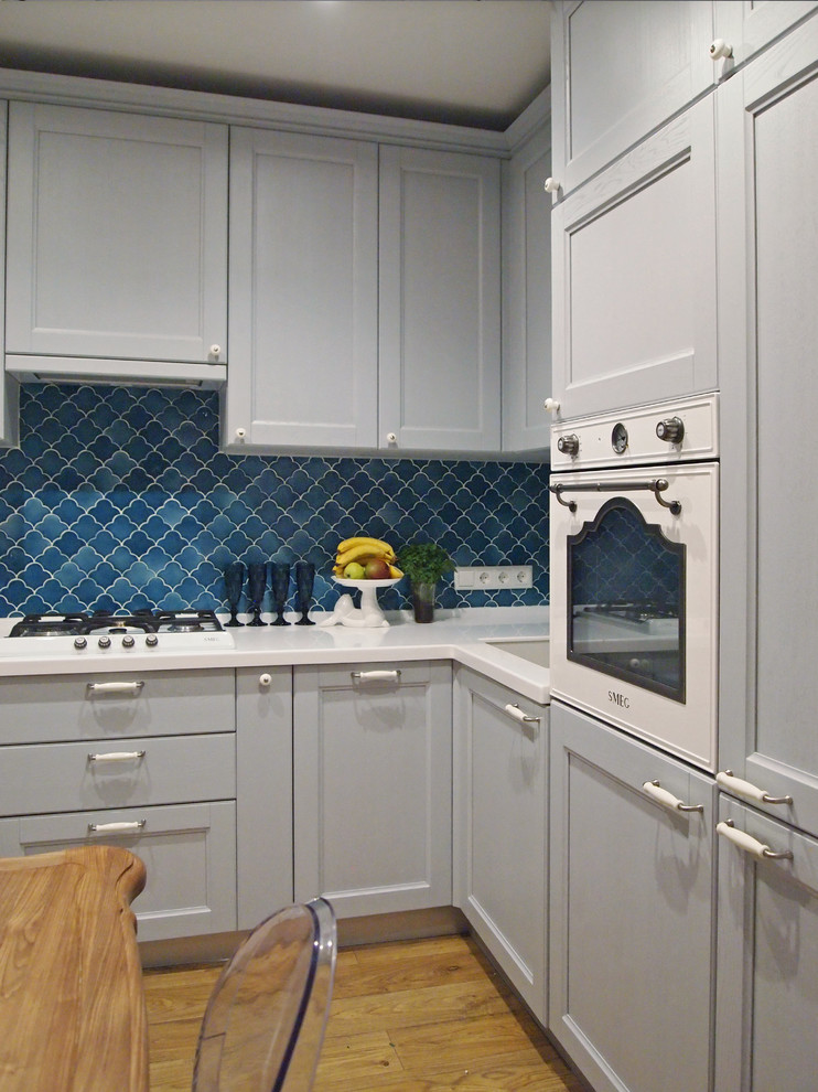 This is an example of a small scandi l-shaped kitchen/diner in Moscow with recessed-panel cabinets, grey cabinets, composite countertops, blue splashback, ceramic splashback, white appliances, laminate floors, no island, a submerged sink and brown floors.