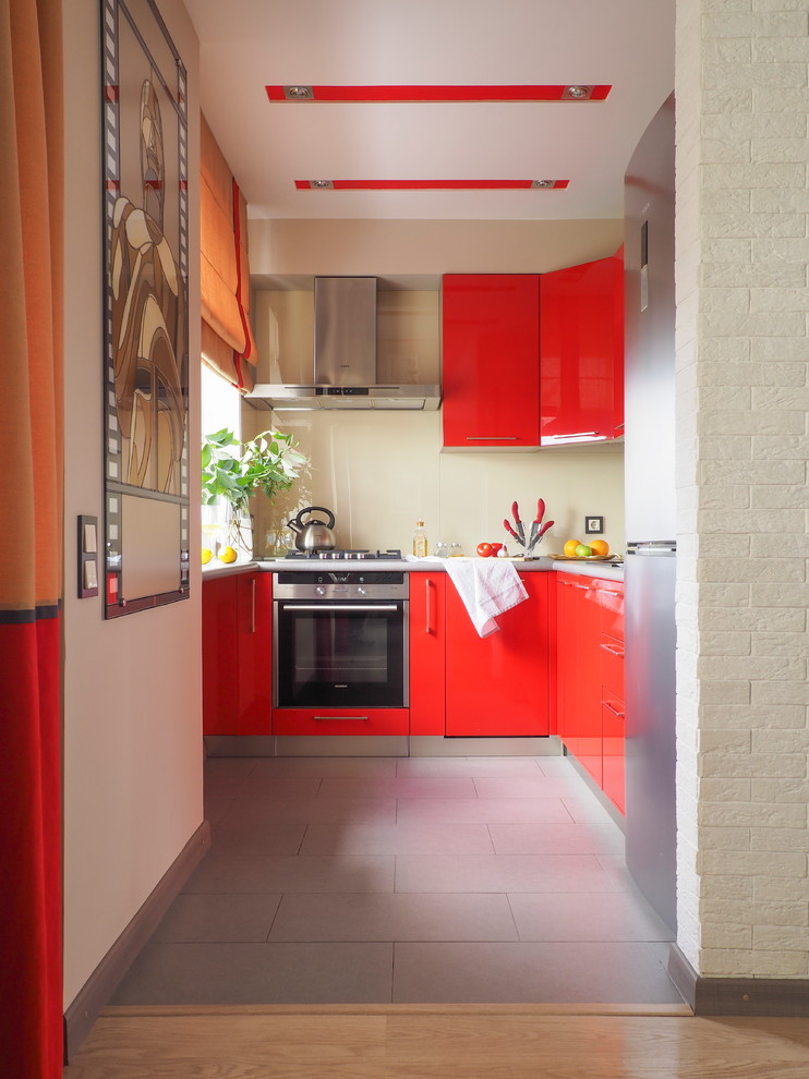 На фото: п-образная кухня в современном стиле с плоскими фасадами, красными фасадами, техникой из нержавеющей стали, серым полом, бежевым фартуком и фартуком из стекла без острова с