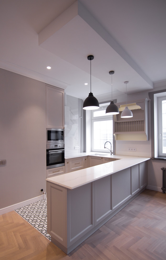 This is an example of a large traditional u-shaped open plan kitchen in Moscow with a double-bowl sink, recessed-panel cabinets, grey cabinets, engineered stone countertops, white splashback, stainless steel appliances, light hardwood flooring, a breakfast bar, brown floors and white worktops.