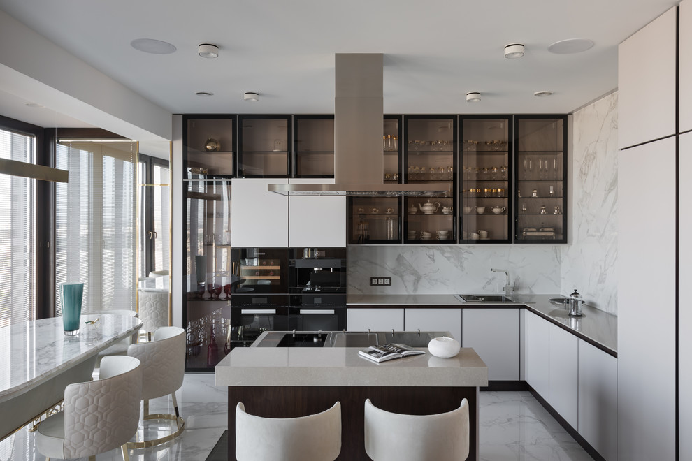 На фото: угловая кухня в современном стиле с накладной мойкой, стеклянными фасадами, белым фартуком, черной техникой, островом и белым полом