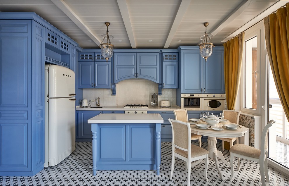 Exemple d'une cuisine américaine chic en L avec un évier encastré, un placard avec porte à panneau surélevé, des portes de placard bleues, une crédence blanche, un électroménager blanc, îlot, un sol multicolore et un plan de travail blanc.