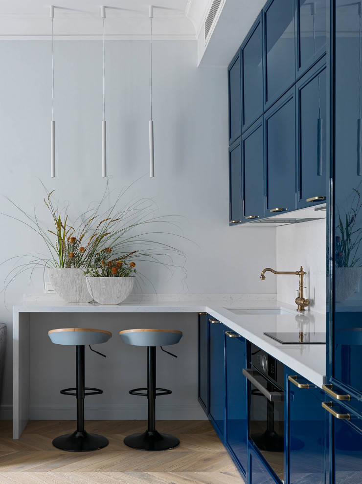 Cette image montre une cuisine design avec un évier encastré, un placard avec porte à panneau encastré, des portes de placard bleues, un électroménager noir, parquet clair, une péninsule et un plan de travail blanc.