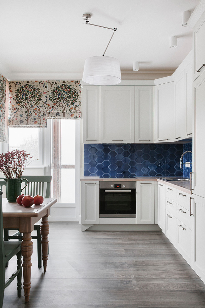 Inredning av ett klassiskt kök, med vita skåp, blått stänkskydd, rostfria vitvaror, en nedsänkt diskho, luckor med profilerade fronter, träbänkskiva, mörkt trägolv och grått golv