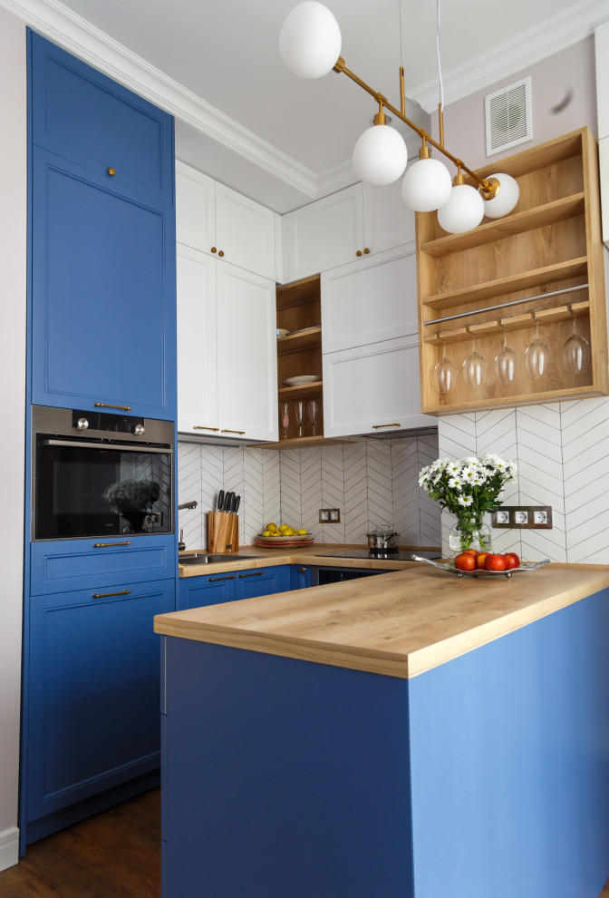 Inredning av ett modernt litet beige beige u-kök, med luckor med infälld panel, blå skåp, träbänkskiva, vitt stänkskydd, rostfria vitvaror, mörkt trägolv, en halv köksö, brunt golv och en nedsänkt diskho