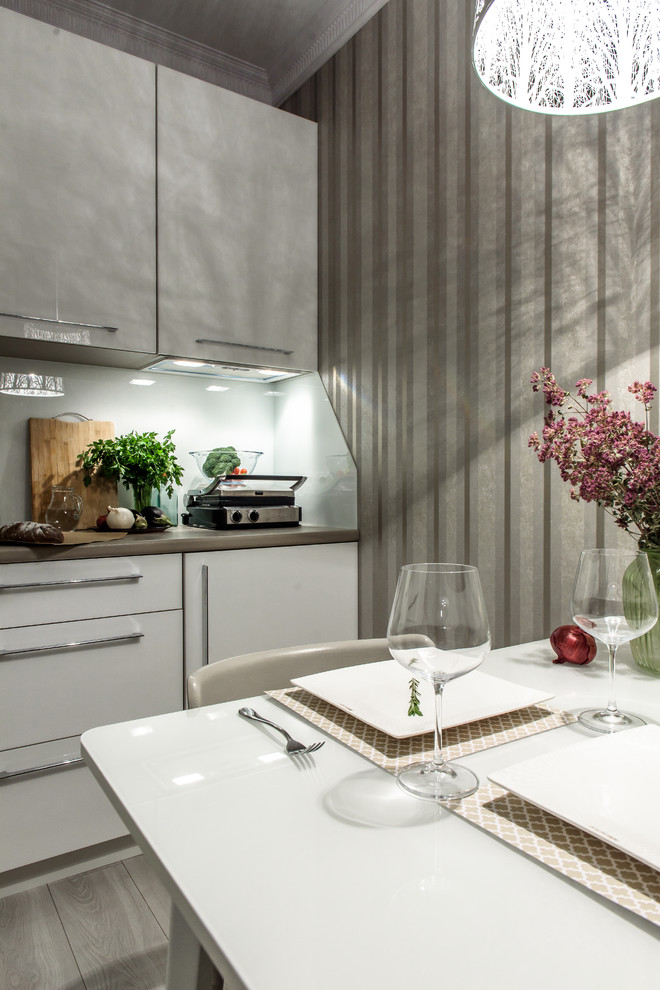 Стильный дизайн: маленькая угловая кухня в современном стиле с обеденным столом, белыми фасадами, белым фартуком, фартуком из стекла, полом из ламината, серым полом и обоями на стенах для на участке и в саду - последний тренд