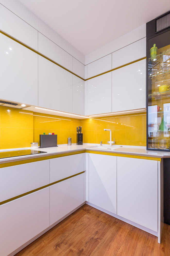 Moderne Küche in L-Form mit Waschbecken, flächenbündigen Schrankfronten, weißen Schränken, Küchenrückwand in Gelb, Glasrückwand, braunem Holzboden und weißer Arbeitsplatte in Sonstige