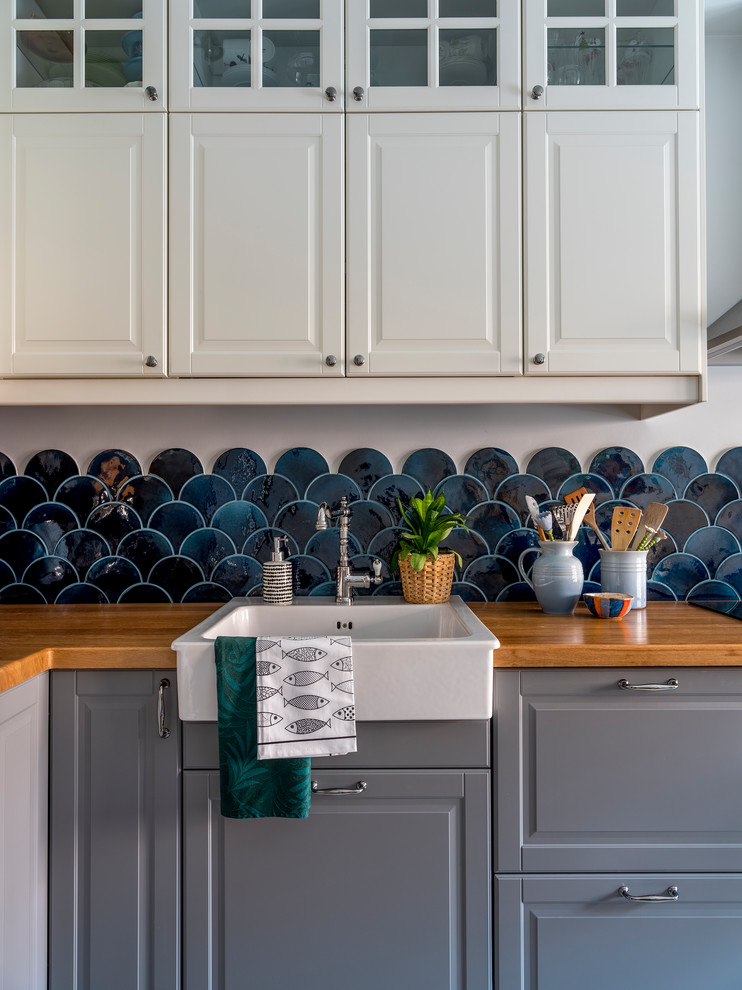 Exemple d'une cuisine bicolore bord de mer en L avec un évier de ferme, un placard avec porte à panneau surélevé, des portes de placard grises, un plan de travail en bois, une crédence bleue et une crédence en céramique.