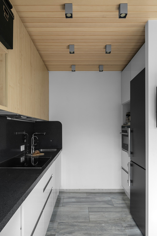 Идея дизайна: узкая параллельная кухня-гостиная в современном стиле с плоскими фасадами, светлыми деревянными фасадами, черным фартуком, техникой из нержавеющей стали и серым полом без острова