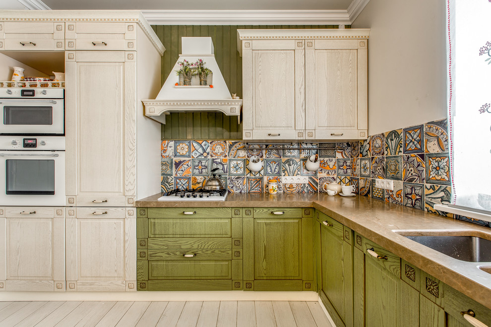 Modelo de cocinas en L campestre con fregadero bajoencimera, puertas de armario verdes, salpicadero multicolor, armarios con paneles con relieve y electrodomésticos blancos