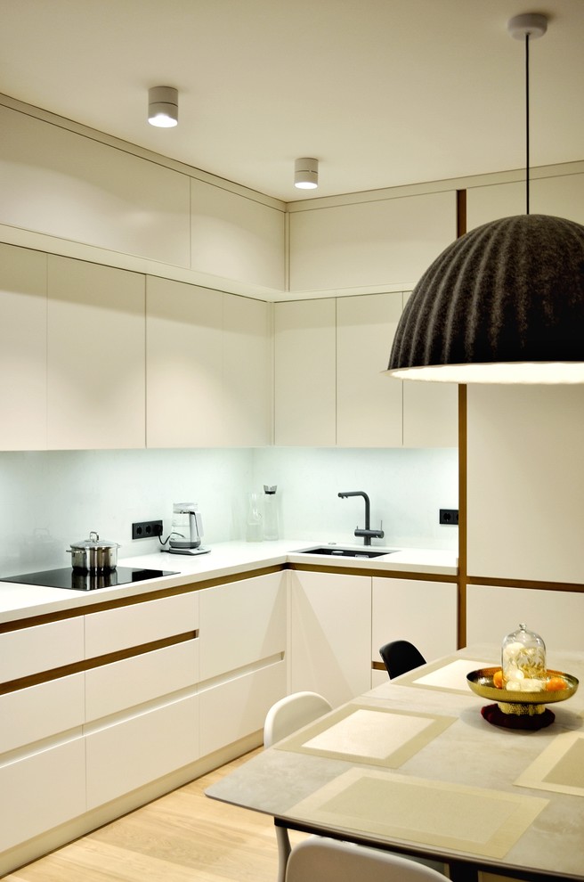 Inspiration for a medium sized contemporary l-shaped open plan kitchen in Moscow with a single-bowl sink, flat-panel cabinets, white cabinets, engineered stone countertops, white splashback, black appliances, light hardwood flooring, no island, brown floors and white worktops.