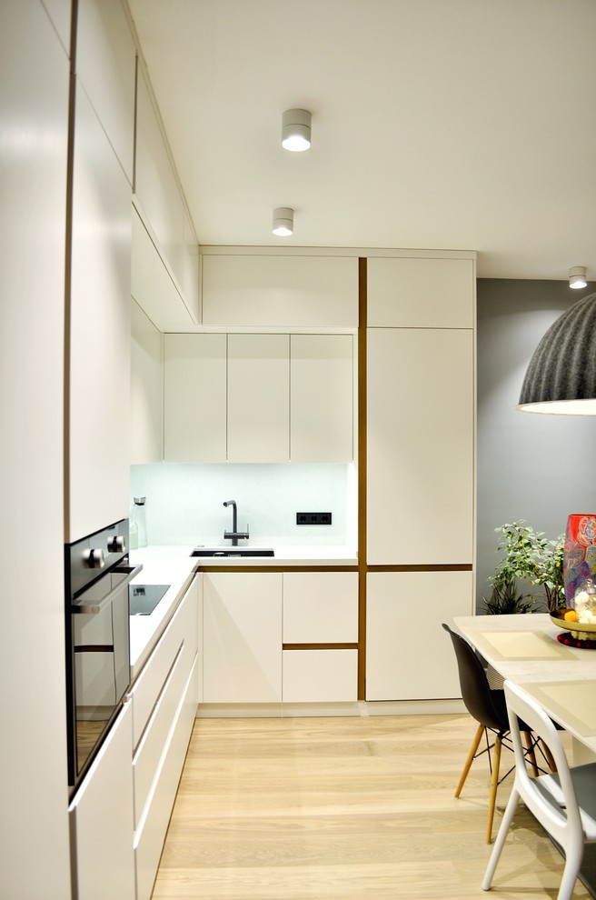 This is an example of a medium sized contemporary l-shaped open plan kitchen in Moscow with a single-bowl sink, flat-panel cabinets, white cabinets, engineered stone countertops, white splashback, black appliances, light hardwood flooring, no island, brown floors and white worktops.