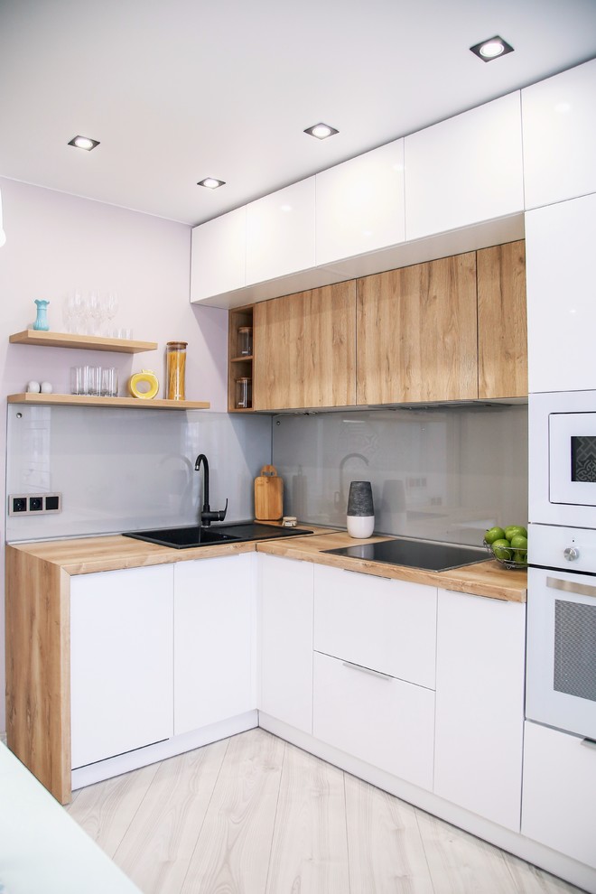 Exempel på ett litet modernt l-kök, med en nedsänkt diskho, släta luckor, vita skåp, träbänkskiva, grått stänkskydd, glaspanel som stänkskydd, vita vitvaror och beiget golv