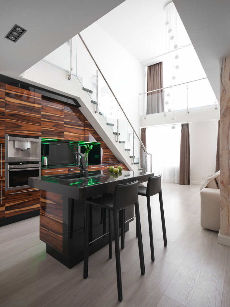 Идея дизайна: параллельная, глянцевая кухня-гостиная в современном стиле с накладной мойкой, плоскими фасадами, фасадами цвета дерева среднего тона, черной техникой, светлым паркетным полом и островом