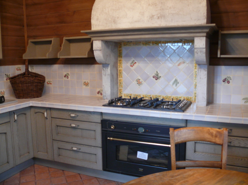 Example of a mid-sized cottage l-shaped terra-cotta tile and red floor eat-in kitchen design in Moscow with a farmhouse sink, recessed-panel cabinets, gray cabinets, tile countertops, white backsplash, black appliances, no island and white countertops