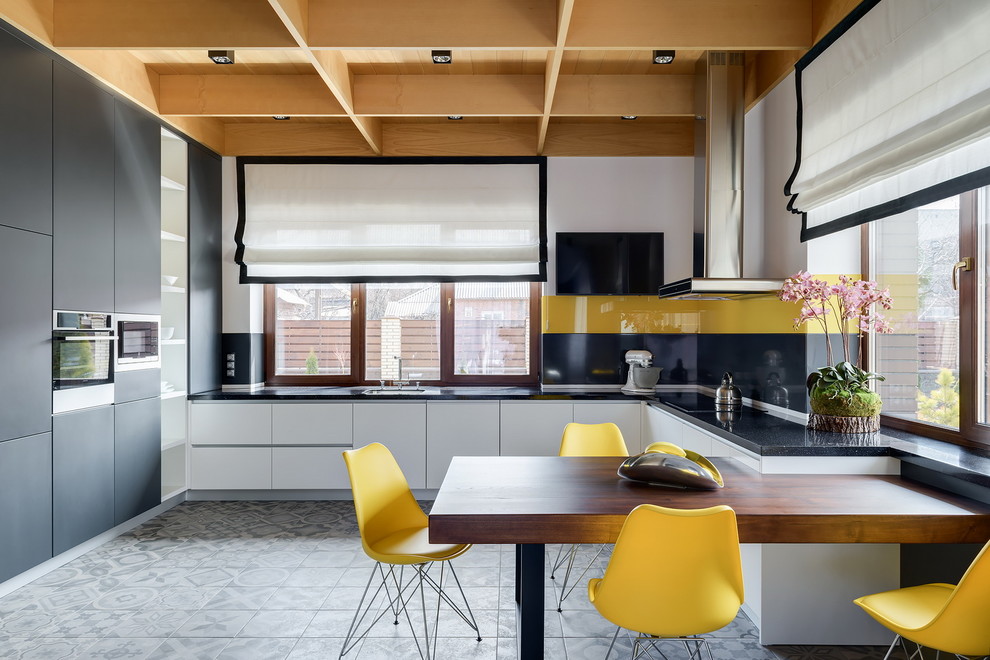 На фото: п-образная кухня в современном стиле с обеденным столом, врезной мойкой, плоскими фасадами, белыми фасадами, желтым фартуком, полуостровом, серым полом, черной столешницей и мойкой у окна с