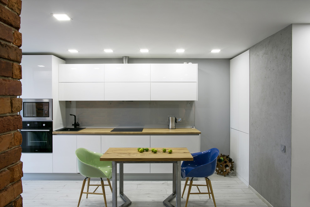 Свежая идея для дизайна: прямая, серо-белая кухня в современном стиле с плоскими фасадами, белыми фасадами, деревянной столешницей, черной техникой, фартуком из стекла и деревянным полом - отличное фото интерьера